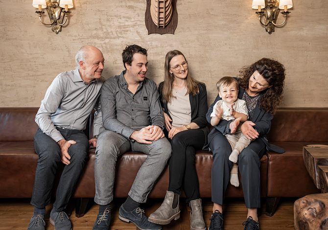 Fünf Personen unterschiedlichen Alters sitzen lächelnd auf einem braunen Ledersofa in einem gemütlichen Raum zusammen.