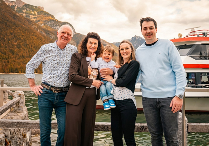 Eine Familie posiert für ein Foto an einem See in der Nähe eines Bootes, wobei der Großvater ein Kleinkind auf dem Arm hält.