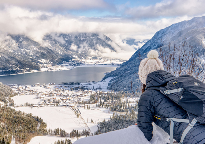 Eine Person in Winterkleidung blickt auf ein verschneites Tal und einen von Bergen umgebenen See.