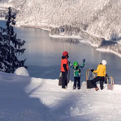 Drei Personen mit Schlitten stehen bei einem Winter-Rodelurlaub auf einem verschneiten Hügel oberhalb des Achensees.