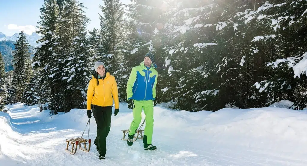 Two people enjoy a winter-tobogganing holiday at Lake Achensee through a snowy pine forest.