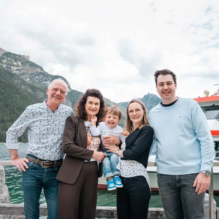 Fünf Personen, darunter ein Kind, posieren und lächeln an einem See mit Bergen und einem Boot im Hintergrund.
